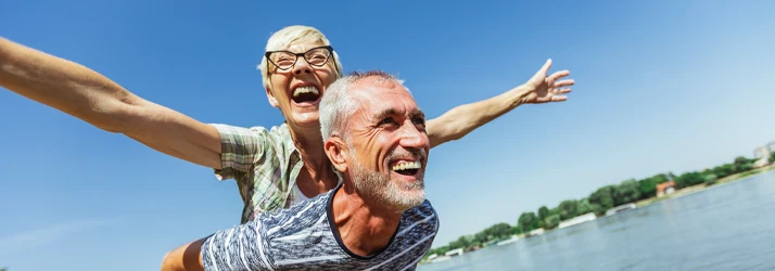Two people near a body of water, with arms extended playfully, under a clear blue sky, evoking a sense of leisure and freedom.